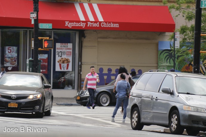 A car turns into Third Avenue from East 106th Street, while a young man carries coffee while crossing the street.